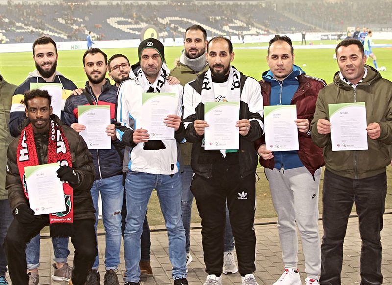 Gruppe von Männern und einer Frau posieren auf einem Fußballfeld mit Urkunden in den Händen. Einige tragen Schals und Jacken mit Fußballmotiven. Im Hintergrund ist das Spielfeld und die Stadiontribüne erkennbar.