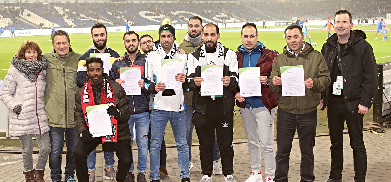 Gruppe von Männern und einer Frau posieren auf einem Fußballfeld mit Urkunden in den Händen. Einige tragen Schals und Jacken mit Fußballmotiven. Im Hintergrund ist das Spielfeld und die Stadiontribüne erkennbar.