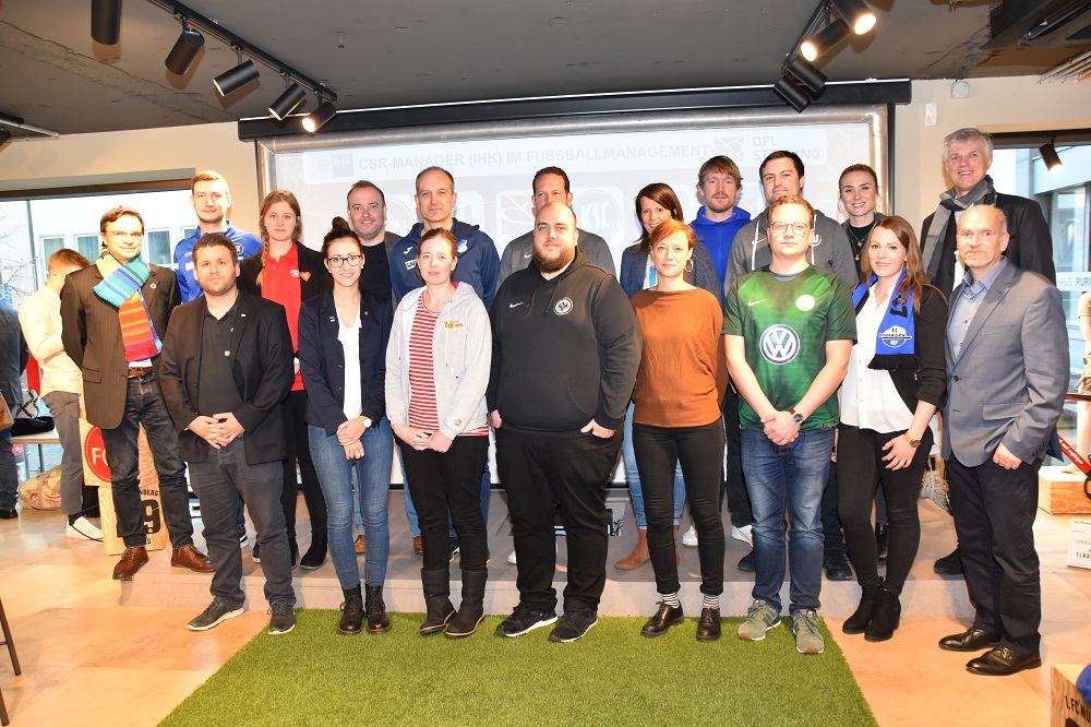 Eine Gruppe von ca. 20 Personen steht lächelnd für ein Gruppenfoto zusammen. Die Teilnehmer der Fortbildung tragen verschiedene Kleidungsstile, von formeller bis leger. Im Hintergrund ist ein Fenster mit Tageslicht zu sehen.
