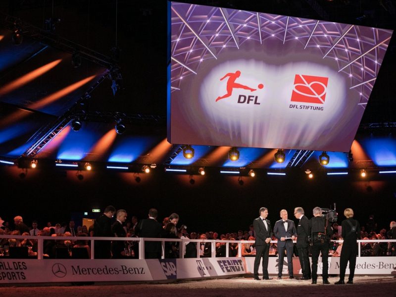 50. Ball des Sports: Auf einer Bühne stehen mehrere Männer in festlicher Abendgarderobe. Im Hintergrund eine große Leinwand mit dem Logo der DFL Stiftung und dem Schriftzug "50. Ball des Sports".