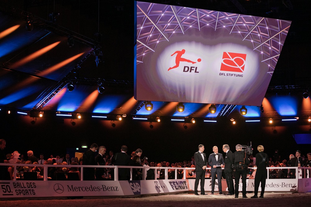 50. Ball des Sports: Auf einer Bühne stehen mehrere Männer in festlicher Abendgarderobe. Im Hintergrund eine große Leinwand mit dem Logo der DFL Stiftung und dem Schriftzug "50. Ball des Sports".