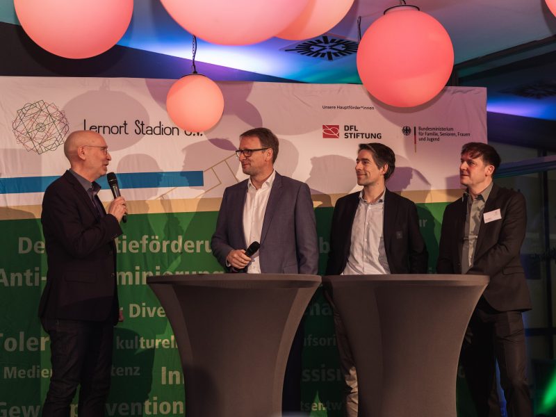 Drei Männer stehen auf einer Bühne vor einer Wand mit dem Schriftzug "DFL Stiftung" und dem Logo des Bundesministeriums für Familie, Senioren, Frauen und Jugend. Einer der Männer hält ein Mikrofon und spricht, während die anderen beiden zuhören. Im Hintergrund sind abstrakte Formen und farbige Elemente zu sehen.