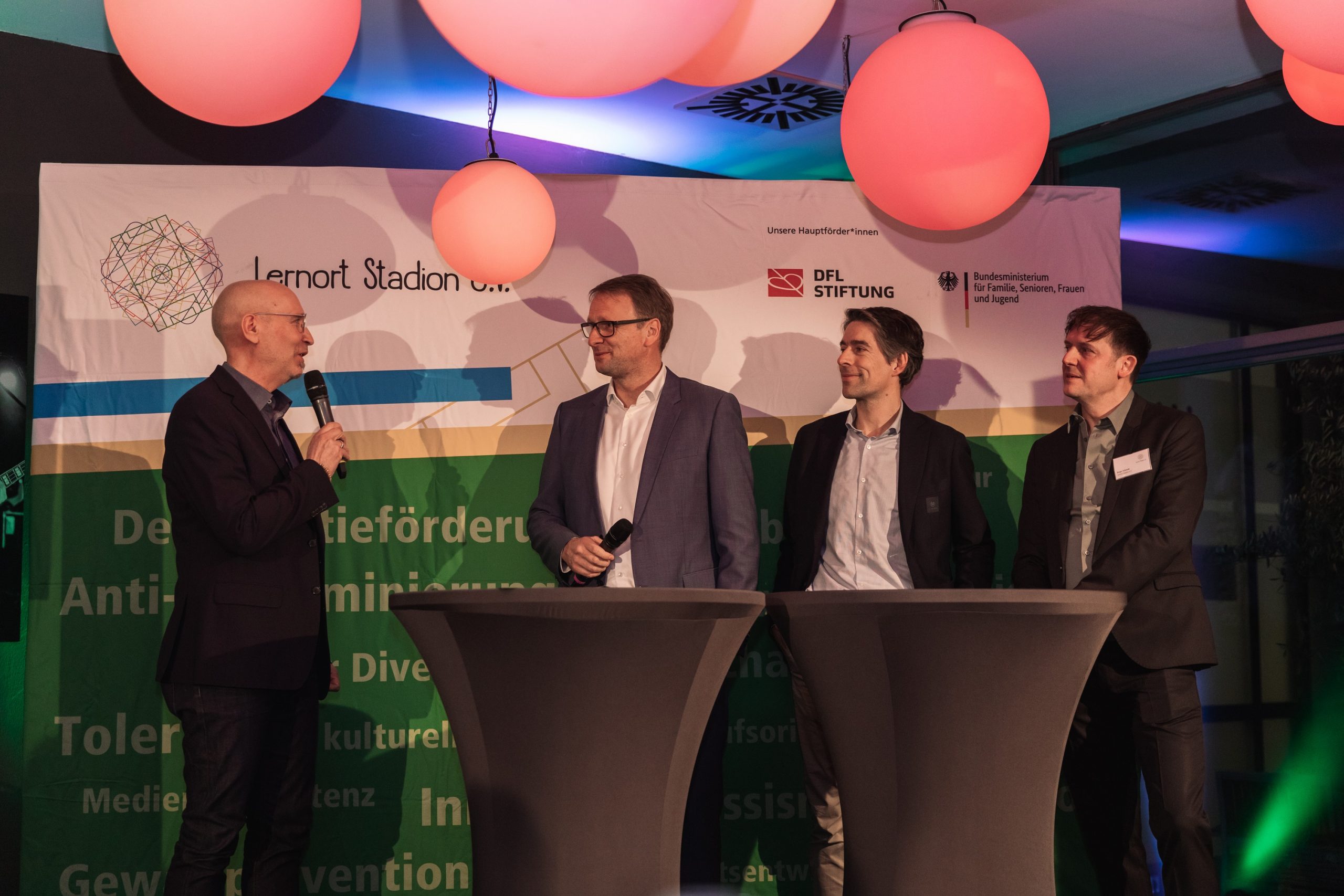 Drei Männer stehen auf einer Bühne vor einer Wand mit dem Schriftzug "DFL Stiftung" und dem Logo des Bundesministeriums für Familie, Senioren, Frauen und Jugend. Einer der Männer hält ein Mikrofon und spricht, während die anderen beiden zuhören. Im Hintergrund sind abstrakte Formen und farbige Elemente zu sehen.