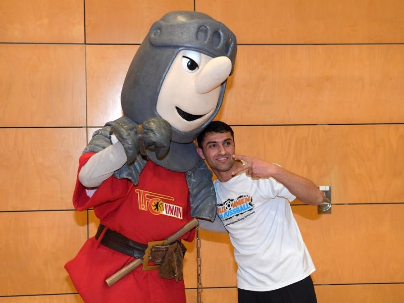 Ein Mann steht mit einem Maskottchen in Ritterrüstung vor einer orangefarbenen Wand. Das Maskottchen, gekleidet in rot-weiß, umarmt den Mann und zeigt den Daumen nach oben. Der Mann trägt ein weißes T-Shirt mit dem Aufdruck "Willkommen im Fußball".