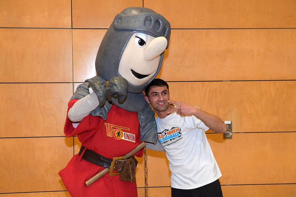 Ein Mann steht mit einem Maskottchen in Ritterrüstung vor einer orangefarbenen Wand. Das Maskottchen, gekleidet in rot-weiß, umarmt den Mann und zeigt den Daumen nach oben. Der Mann trägt ein weißes T-Shirt mit dem Aufdruck "Willkommen im Fußball".