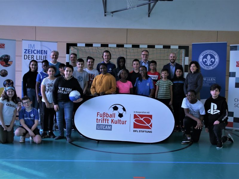 Eine Gruppe von Kindern und Jugendlichen steht in einer Turnhalle vor einer Wand mit Bannern. Im Vordergrund steht ein Banner mit der Aufschrift "Fußball trifft Kultur - LITCAMI" und dem Logo der DFL Stiftung. Die Gruppe besteht aus einer vielfältigen Mischung von Kindern und Jugendlichen unterschiedlicher Herkunft. Einige halten Fußbälle, andere lächeln in die Kamera. Im Hintergrund sind weitere Banner und ein Basketballkorb zu sehen.