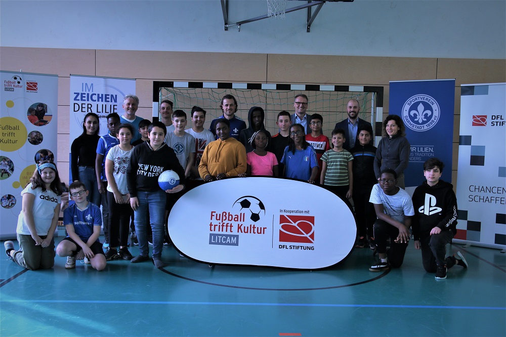Eine Gruppe von Kindern und Jugendlichen steht in einer Turnhalle vor einer Wand mit Bannern. Im Vordergrund steht ein Banner mit der Aufschrift "Fußball trifft Kultur - LITCAMI" und dem Logo der DFL Stiftung. Die Gruppe besteht aus einer vielfältigen Mischung von Kindern und Jugendlichen unterschiedlicher Herkunft. Einige halten Fußbälle, andere lächeln in die Kamera. Im Hintergrund sind weitere Banner und ein Basketballkorb zu sehen.