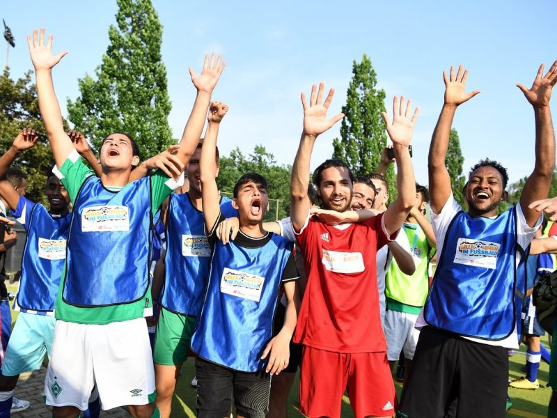 Jubelnde Teilnehmer eines Fußballtrainings, die mit erhobenen Armen ihre Freude zeigen. Einige tragen blaue Trainingswesten, andere ein rotes Trikot und grüne Shorts. Im Hintergrund sind weitere Personen und ein Fußballfeld zu erkennen.