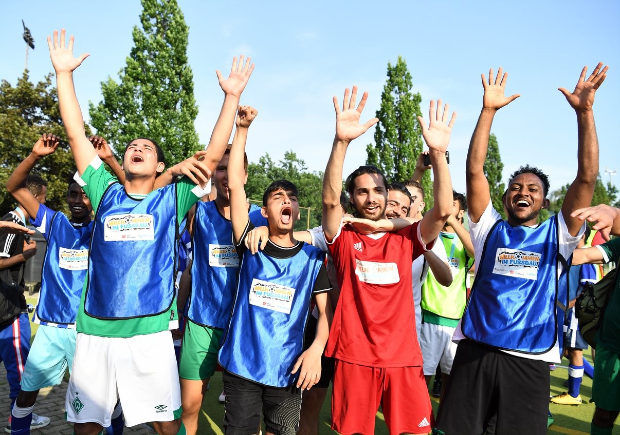 Jubelnde Teilnehmer eines Fußballtrainings, die mit erhobenen Armen ihre Freude zeigen. Einige tragen blaue Trainingswesten, andere ein rotes Trikot und grüne Shorts. Im Hintergrund sind weitere Personen und ein Fußballfeld zu erkennen.