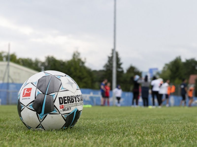 Ein Fußball liegt auf dem grünen Rasen eines Fußballfeldes. Im Hintergrund sind verschwommene Personen zu sehen, die an einem Workshop teilnehmen. Im Vordergrund ist der Schriftzug "DERBYSTAR" und das Logo der "Bundesliga" auf dem Ball erkennbar.