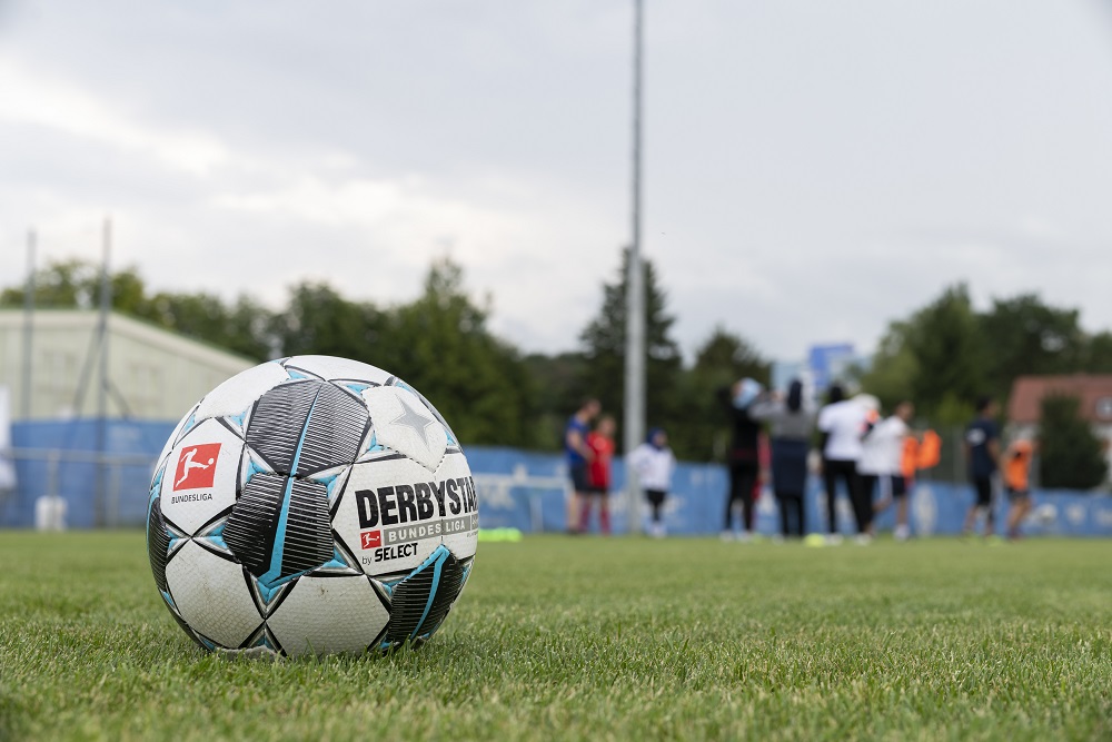 Ein Fußball liegt auf dem grünen Rasen eines Fußballfeldes. Im Hintergrund sind verschwommene Personen zu sehen, die an einem Workshop teilnehmen. Im Vordergrund ist der Schriftzug "DERBYSTAR" und das Logo der "Bundesliga" auf dem Ball erkennbar.
