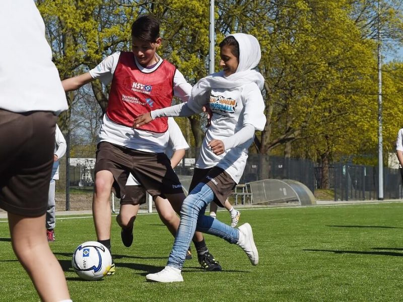 Fünf junge Menschen spielen auf einem Fußballfeld, darunter ein Kind mit Kopftuch, während es einen Ball spielt.