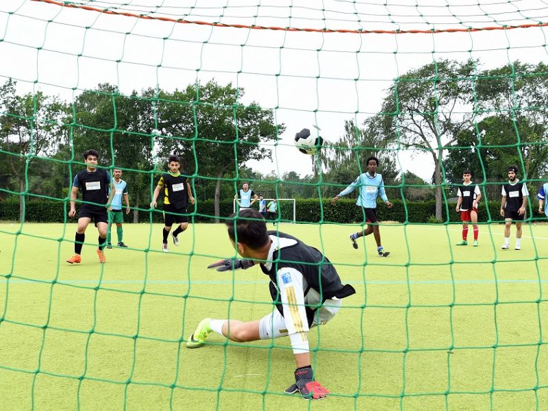 Ein Fußballtorwart springt nach einem Ball, der in Richtung Tor fliegt, während andere Spieler auf dem grünen Kunstrasenplatz stehen. Im Hintergrund sind Bäume und der Himmel zu sehen.