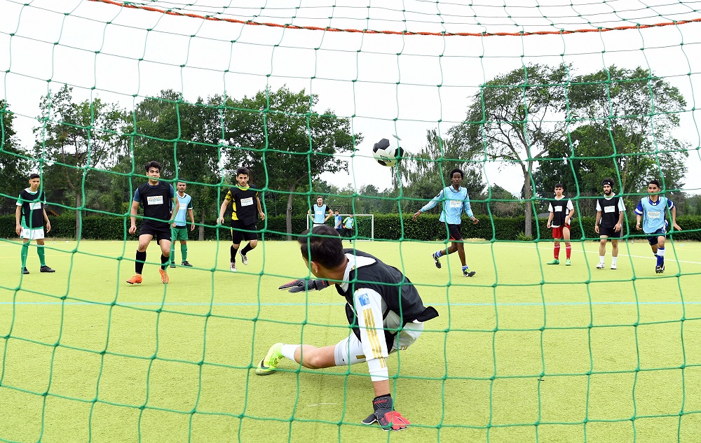 Ein Fußballtorwart springt nach einem Ball, der in Richtung Tor fliegt, während andere Spieler auf dem grünen Kunstrasenplatz stehen. Im Hintergrund sind Bäume und der Himmel zu sehen.