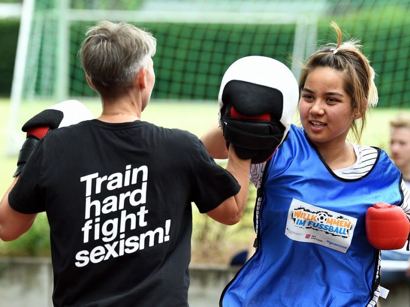 Zwei Fußballspielerinnen trainieren im Freien. Die Spielerin links, mit dem Rücken zum Betrachter, trägt ein T-Shirt mit der Aufschrift "Train hard fight sexism!". Die Spielerin rechts trägt ein blaues Trikot mit dem Logo "Willkommen im Fußball" und Boxhandschuhe.