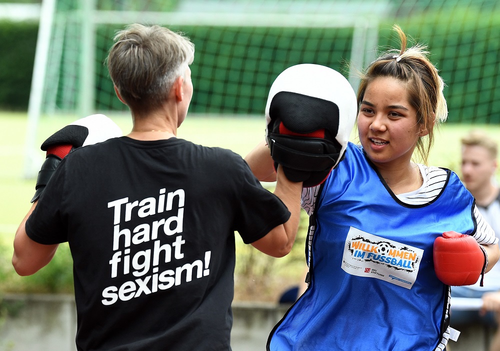 Zwei Fußballspielerinnen trainieren im Freien. Die Spielerin links, mit dem Rücken zum Betrachter, trägt ein T-Shirt mit der Aufschrift "Train hard fight sexism!". Die Spielerin rechts trägt ein blaues Trikot mit dem Logo "Willkommen im Fußball" und Boxhandschuhe.