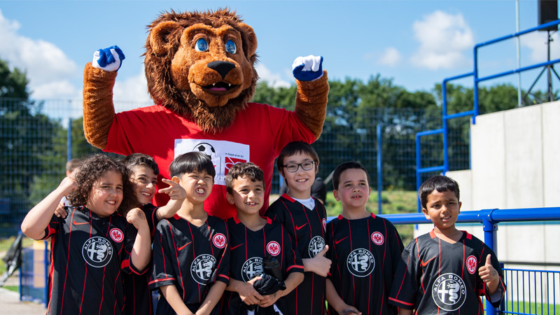 Sieben Jungs in schwarzen Trikots von Eintracht Frankfurt stehen vor Litti, dem Maskottchen von "Fußball trifft Kultur". Litti ist ein Löwe mit rotem T-Shirt.