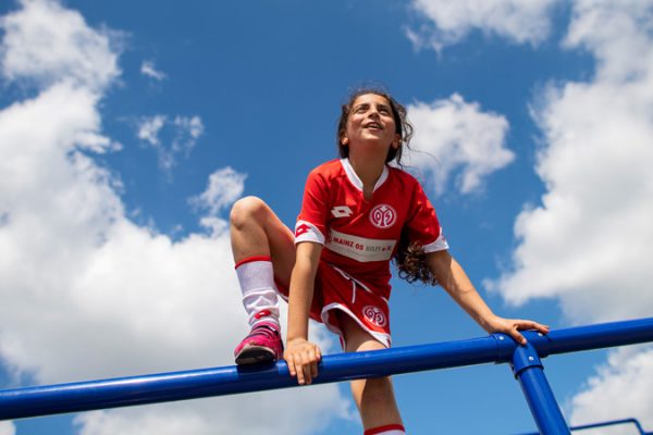 Ein Mädchen mit rotem Fußballtrikot klettert auf ein Geländer. Ihr rechter Fuß und ihre Hände sind auf dem Geländer, das link Bein hängt herunter.