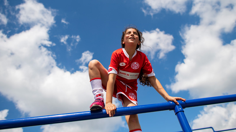 Ein Mädchen mit rotem Fußballtrikot klettert auf ein Geländer. Ihr rechter Fuß und ihre Hände sind auf dem Geländer, das link Bein hängt herunter.