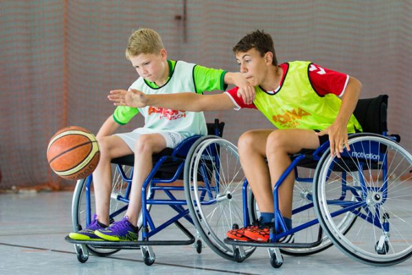 Spielszene vom Rollstuhl-Basketball: Ein Junge im gelben Leibchen streckt sich im Rollstuhl sitzend nach dem Ball. Sein ebenso im Rollstuhl sitzender Gegner, ein Junge im weißen Leibchen, packt ihm dabei auf die Schulter.
