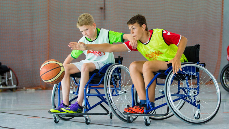 Spielszene vom Rollstuhl-Basketball: Ein Junge im gelben Leibchen streckt sich im Rollstuhl sitzend nach dem Ball. Sein ebenso im Rollstuhl sitzender Gegner, ein Junge im weißen Leibchen, packt ihm dabei auf die Schulter.