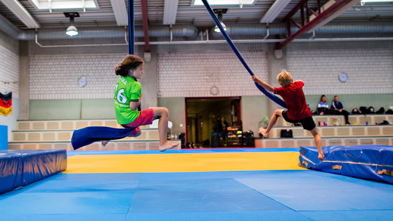 Zwei Kinder springen in einer Turnhalle an Ringen, während sie an einem Seil schwingen. Im Hintergrund sind weitere Kinder und eine Zuschauertribüne zu sehen.