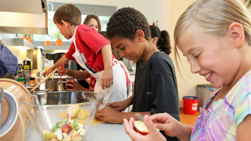 Eine Gruppe von Kindern steht in einer Küche um eine Arbeitsplatte herum und bereitet gemeinsam Obstsalat zu. Ein Kind schält einen Apfel, während andere bereits geschnittenes Obst in einer großen Schüssel vermischen.