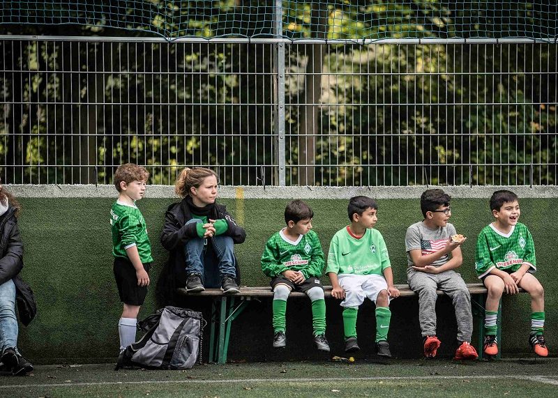 Junge Fußballspielerinnen und Fußballspieler des Bremer „Willkommen im Fußball“-Bündnisses sitzen auf den Bänken neben dem Fußballfeld und schauen das Spiel zu.