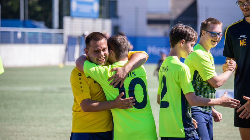 Im Vordergrund umarmen sich zwei Fußballspieler mit gelben Trikots. Im Hintergrund geben sich drei weitere Jungs die Hände.
