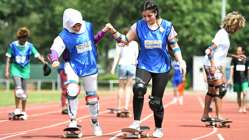 Zwei junge Frauen mit Migrationshintergrund fahren Skateboard. Dabei halten sie sich an jeweils einer Hand fest.