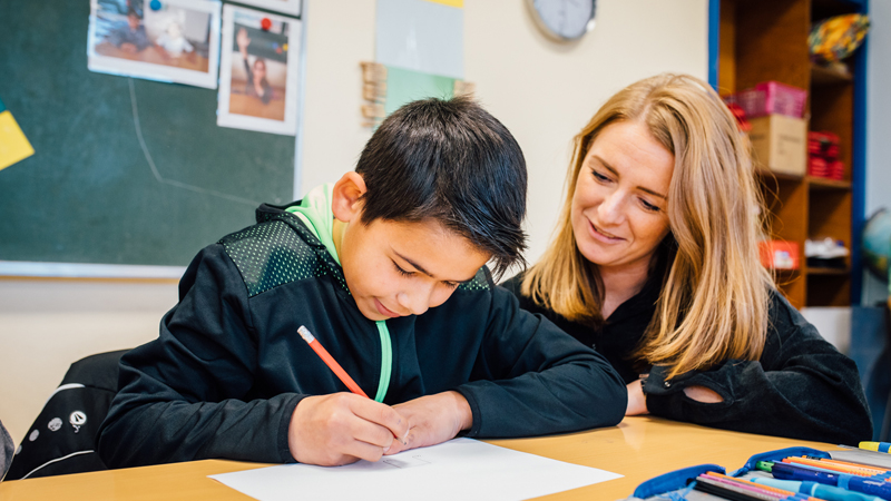 Ein Junge sitzt am Schultisch und schreibt etwas. Eine Frau schaut ihm über die Schulter zu.