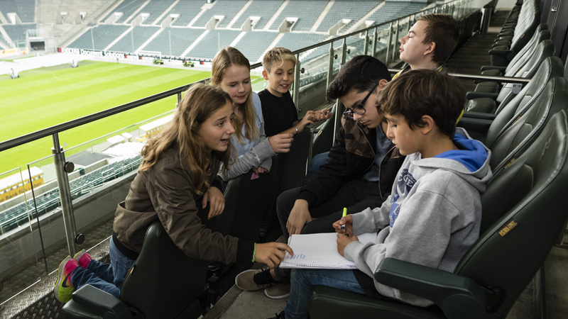 Eine Gruppe Kinder und Jugendlicher Teilnehmender des Programms Lernort Stadion sitzt auf den leeren Tribünen eines Fußballstadions. Ein Mädchen zeigt einem jungen etwas auf einem Blatt Papier, dass dieser in der Hand hält.