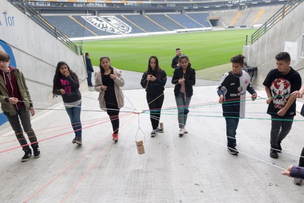 A group of young people participating in the "Lernort Stadion" program stands in a semicircle at one of the gates of a soccer stadium. The playing field can be seen in the background. Together, the young people try to move an object in the middle with the help of ropes.