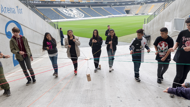 A group of young people participating in the "Lernort Stadion" program stands in a semicircle at one of the gates of a soccer stadium. The playing field can be seen in the background. Together, the young people try to move an object in the middle with the help of ropes.