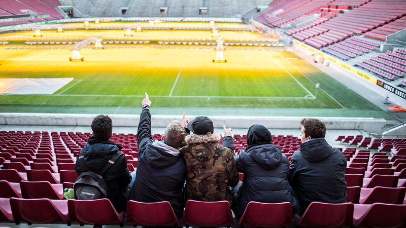 A small group of students sits together in an empty soccer stadium and looks out onto the field from the stands. The group is taking part in the DFL Foundation's 