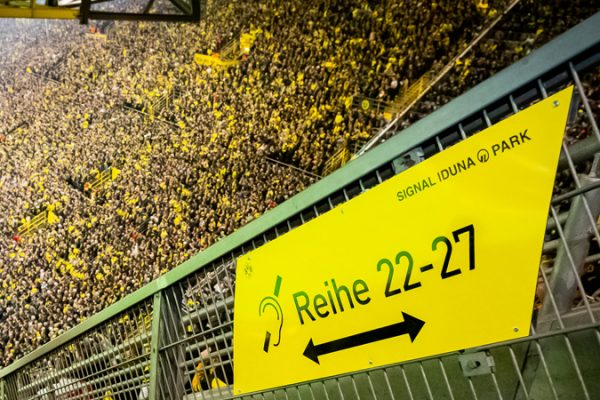 An einem Geländer im Dortmunder Signal Iduna Park hängt ein gelbes Schild. Auf diesem Schild ist in Schwarz folgendes zu sehen: das internationale Symbol für Hörschädigung (ein Ohr mit Strich) sowie Reihe 22 - 27.