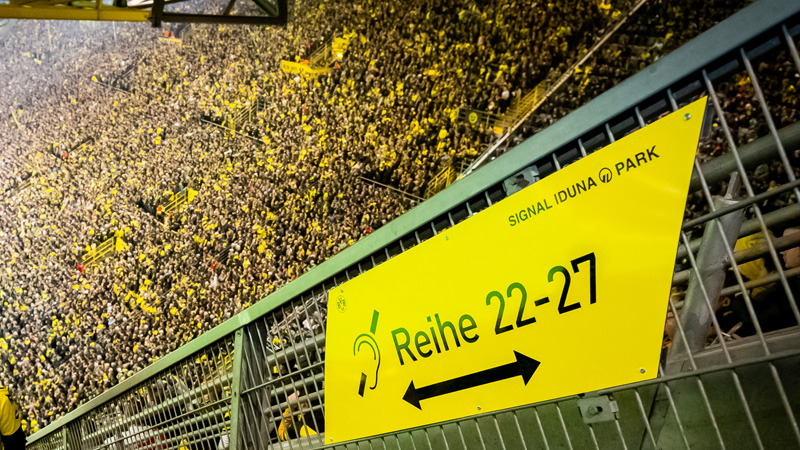 An einem Geländer im Dortmunder Signal Iduna Park hängt ein gelbes Schild. Auf diesem Schild ist in Schwarz folgendes zu sehen: das internationale Symbol für Hörschädigung (ein Ohr mit Strich) sowie Reihe 22 - 27.