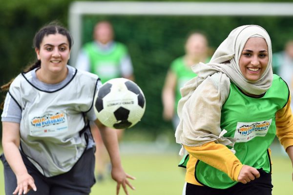 Zwei jugendliche Mädchen mit Migrationshintergrund, die am Programm Willkommen im Fußball teilnehmen, laufen einem Ball hinterher.