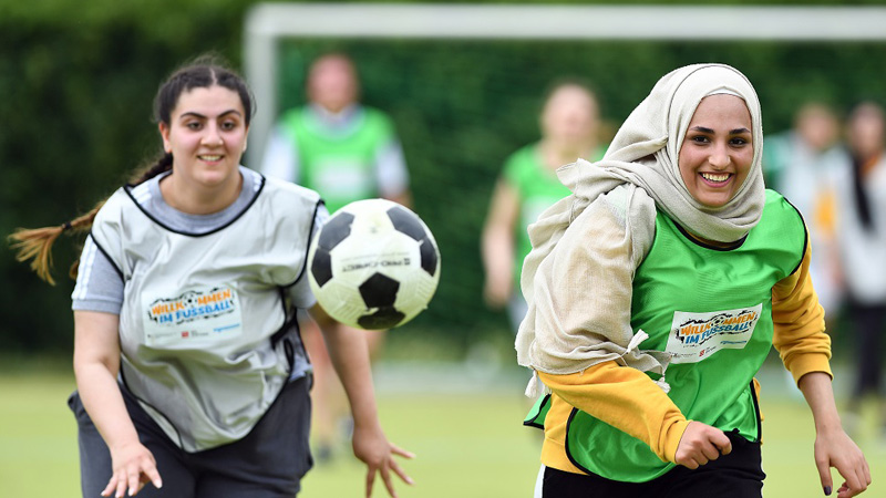 Zwei jugendliche Mädchen mit Migrationshintergrund, die am Programm Willkommen im Fußball teilnehmen, laufen einem Ball hinterher.