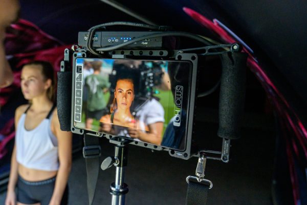 Eine junge Fußballspielerin wird während eines Interviews gefilmt. Im Vordergrund ist ein Monitor zu sehen, auf dem ihr Gesicht zu erkennen ist, während im Hintergrund eine zweite Spielerin steht.