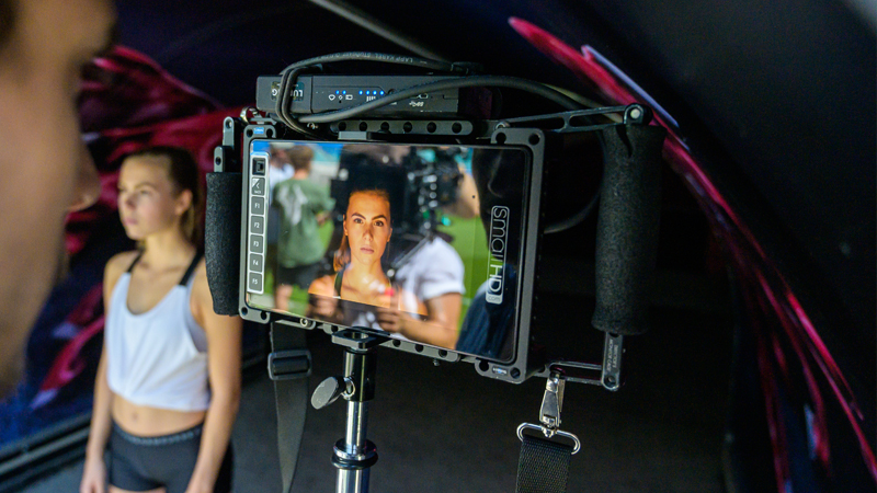 Eine junge Fußballspielerin wird während eines Interviews gefilmt. Im Vordergrund ist ein Monitor zu sehen, auf dem ihr Gesicht zu erkennen ist, während im Hintergrund eine zweite Spielerin steht.