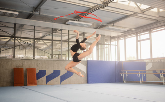 Die rhythmische Sportgymnastin Noemi Peschel demonstriert in einer Turnhalle einen Sprung. In der Hand hält sie ein rotes Schwungband.