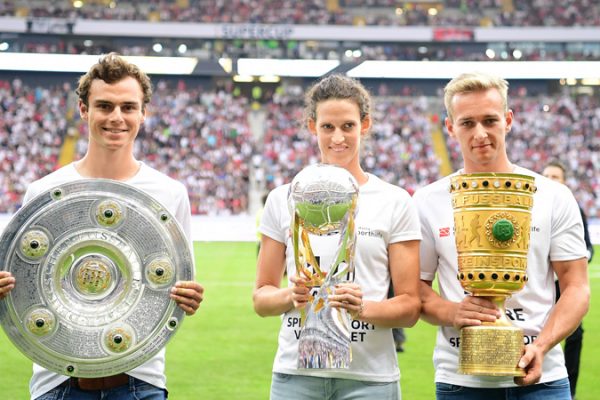 Athletes Vinzenz Geiger, Lena Schöneborn and Johannes Floors stand in a soccer stadium before the kickoff of the Super Cup. Each of them is holding a cup in his hands.