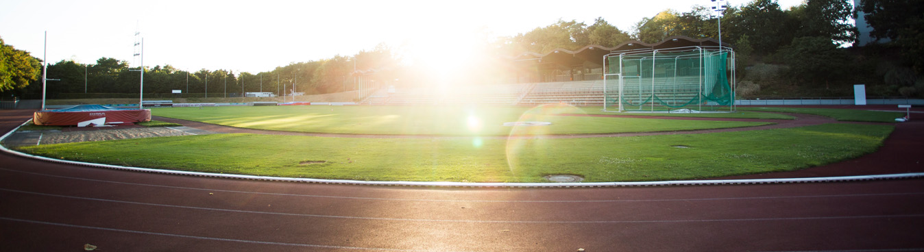 Fußballfeld mit roter Laufbahn, hell erleuchtet von der tiefstehenden Sonne. Im Hintergrund sind grüne Bäume und Tribünen zu erkennen.
