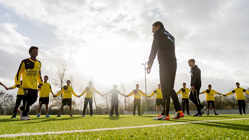 Jugendliche mit Migrationshintergrund stehen Hand in Hand in einem Kreis auf einem Fußballplatz. Sie tragen gelbe Trikots von Borussia Dortmund. Im Vordergrund sind die zwei Trainer der Jugendlichen zu erkennen.