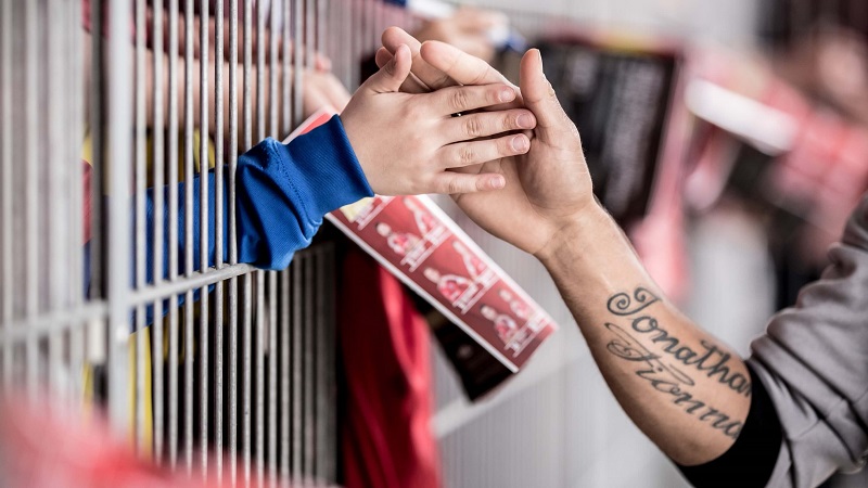 Eine kleine Hand, die durch den Zaun eines Fußballstadions durchgestreckt wird und von einer anderen Hand abgeklatscht wird.
