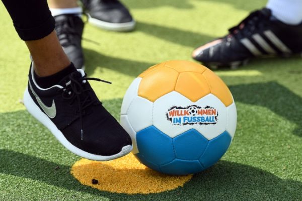 Ein Fuß, der gegen einen Fußball tritt. Der Fußball hat die Farben orange, weiß und gelb, in der Mitte des Balles ist das Logo von Willkommen im Fußball zu sehen.