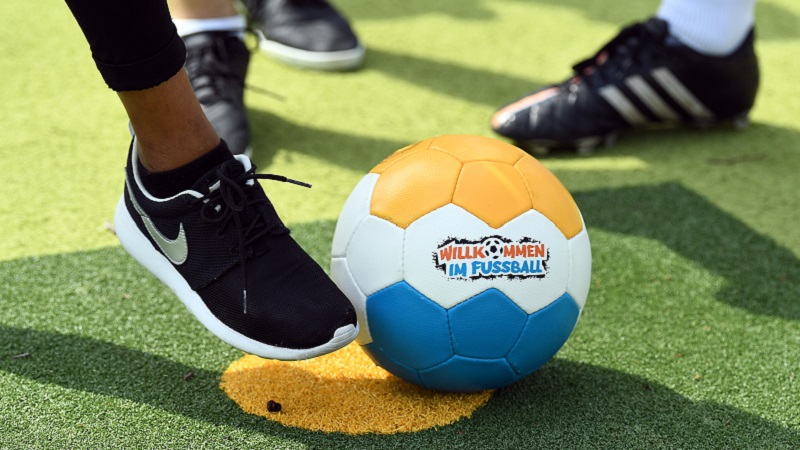 Ein Fuß, der gegen einen Fußball tritt. Der Fußball hat die Farben orange, weiß und gelb, in der Mitte des Balles ist das Logo von Willkommen im Fußball zu sehen.