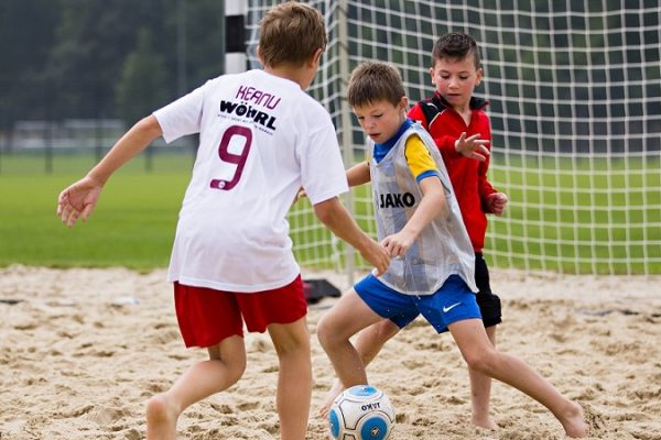 Drei Kinder spielen gemeinsam Fußball im Sand. Im Hintergrund ist das Tor des Beachsoccer-Feldes zu erkennen.
