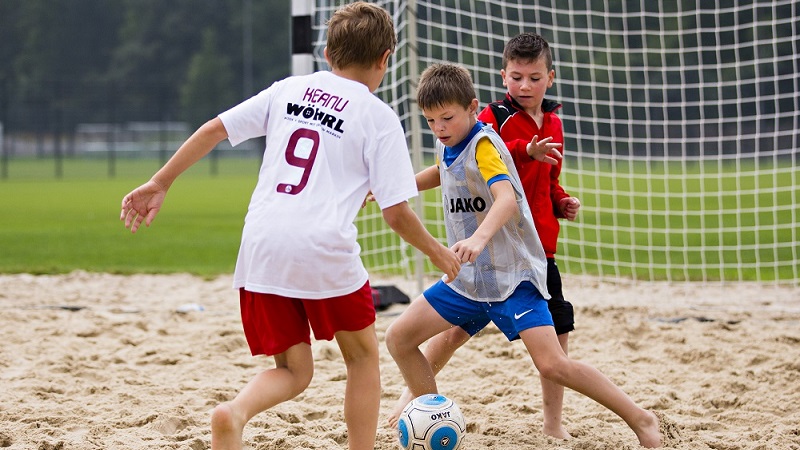 Drei Kinder spielen gemeinsam Fußball im Sand. Im Hintergrund ist das Tor des Beachsoccer-Feldes zu erkennen.
